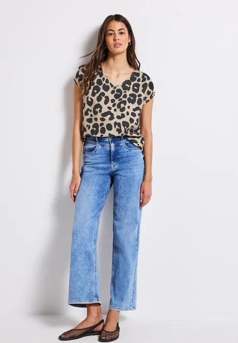 Frau in Leo-T-Shirt, blauer Jeans und schwarzen Schuhen steht vor weißer Wand.